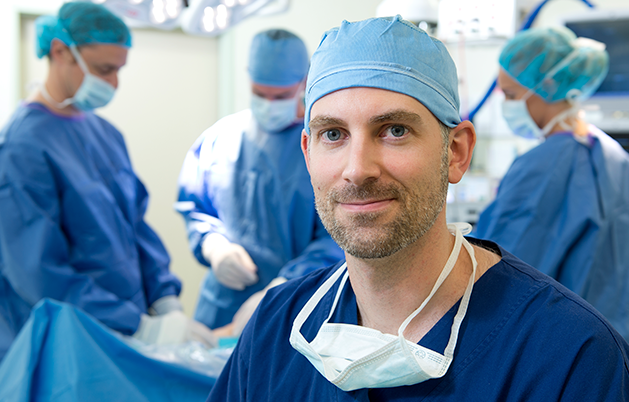 Orthopaedic surgeon in blue surgical scrubs, cap and mask with surgical team preparing in operating theatre background